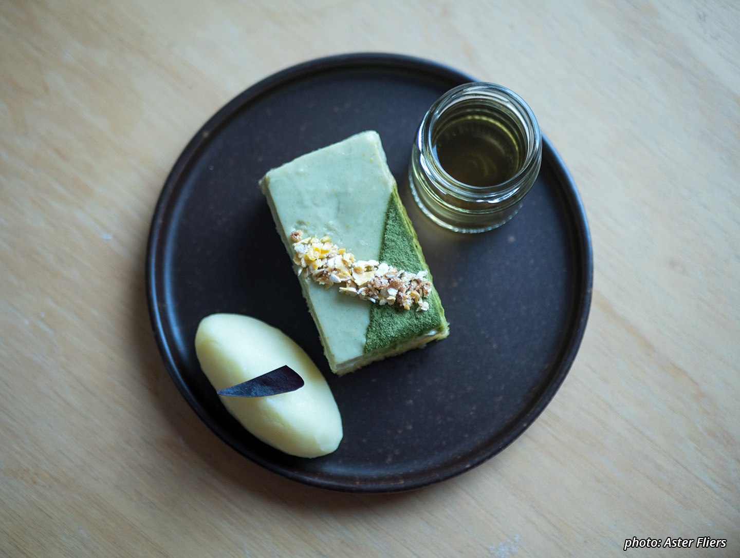 A photograph of an exquisite looking small yellow wheat&corn-based lemon cake with a green (barley)grass&orange soy-custard and crunchy corn-topping, an intense lemon sorbet with a sliver of purple clover and a shot of cornsilk&caraway-tea.