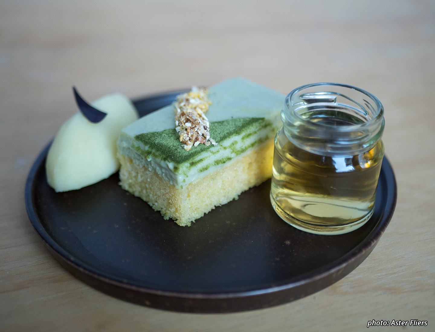 A photograph of an exquisite looking small yellow wheat&corn-based lemon cake with a green (barley)grass&orange soy-custard and crunchy corn-topping, an intense lemon sorbet with a sliver of purple clover and a shot of cornsilk&caraway-tea.