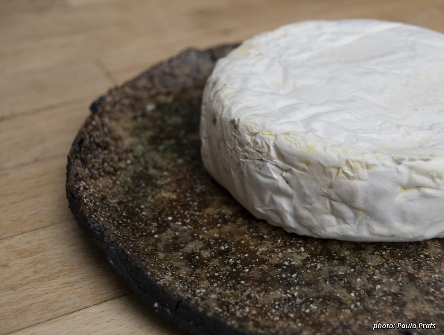 Picture of a black plate textured with sand, on which a beautiful craggly white camembert has been placed.