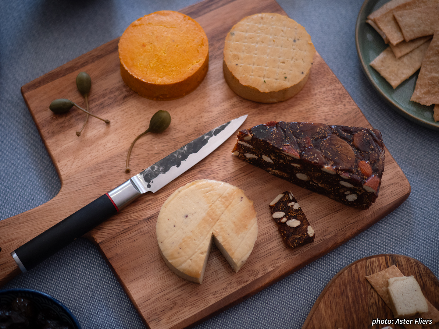 Picture of fermented cashew cheeses, crackers and pickled caperberries on a board with a dried fig loaf. There's aged cashew with miso, aged cashew and hazelnut with annatto, and smoked aged cashew.